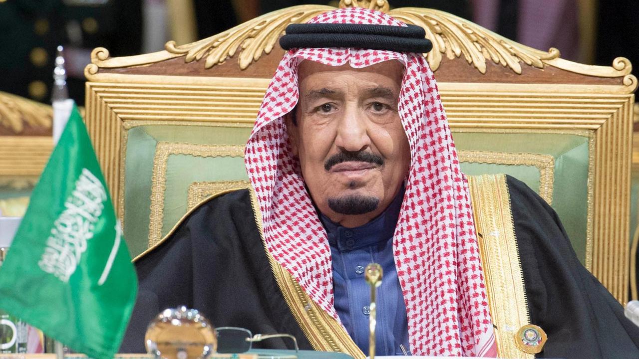 Salman sitzt mit traditionellem Kopftuch auf einer Art Thron an einem Tisch. Neben ihm steht ein kleine Flagge Sausi-Arabiens. Im Hintergrund sieht man unscharf zwei Männer mit dekorierten Uniformen. Salman sitzt mit traditionellem Kopftuch auf einer Art Thron an einem Tisch. Neben ihm steht ein kleine Flagge Sausi-Arabiens. Im Hintergrund sieht man unscharf zwei Männer mit dekorierten Uniformen.