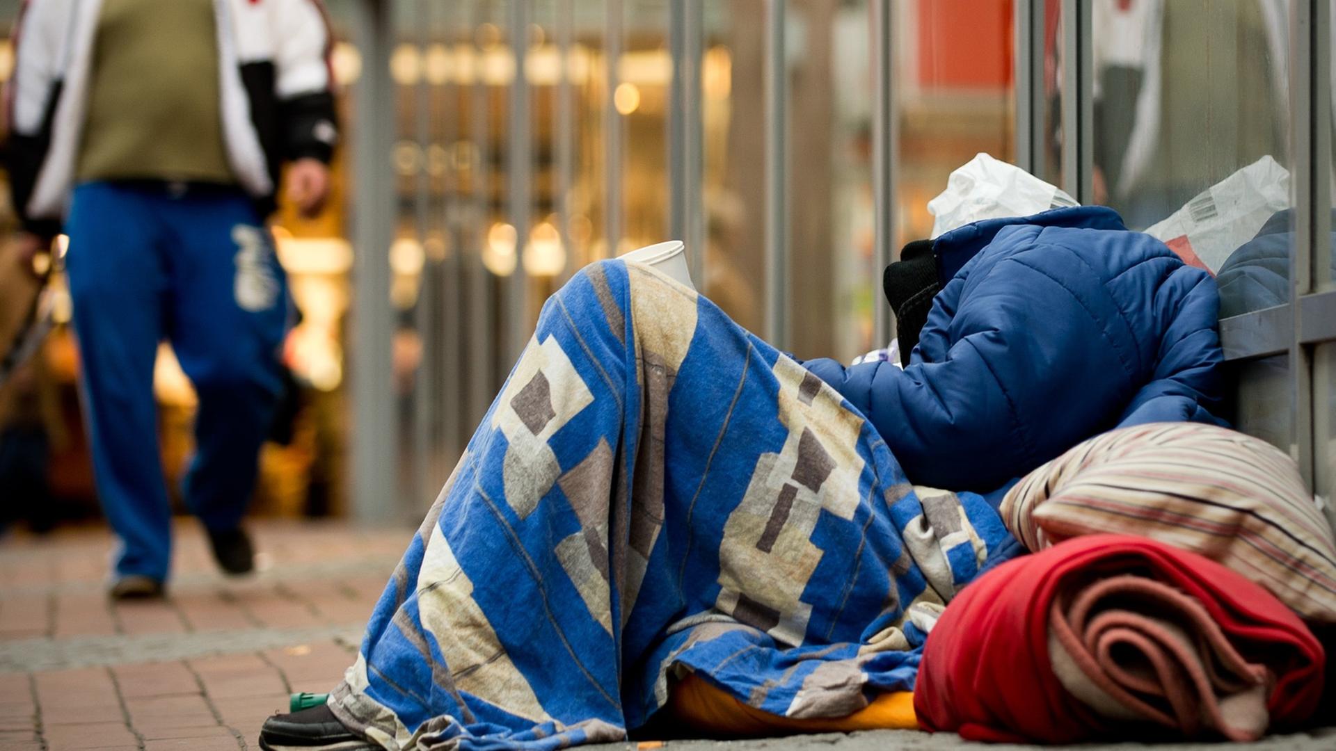 Ein Obdachloser schläft in einer Fußgängerzone in Dortmund, eingepackt in Decken und zwischen Plastiktüten auf dem Boden, während ein Mann vorbei läuft. Ein Obdachloser schläft in einer Fußgängerzone in Dortmund, eingepackt in Decken und zwischen Plastiktüten auf dem Boden, während ein Mann vorbei läuft.
