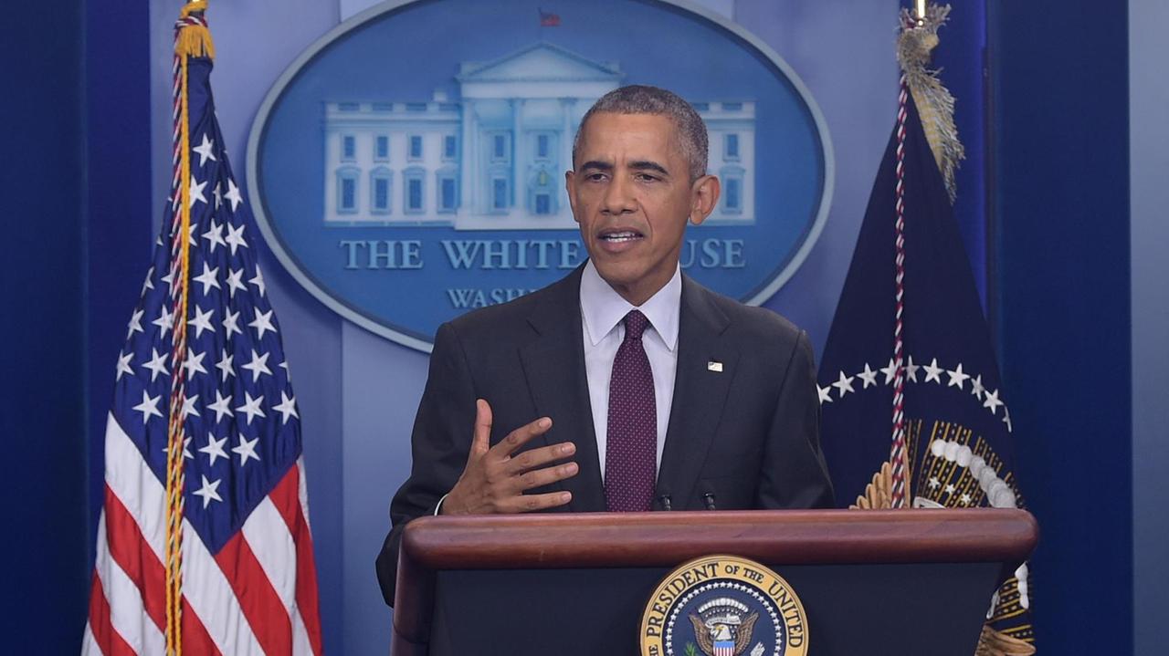 Der Präsident steht hinter dem Pult des Pressekonferenz-Raums vor blauem Hintergrund mit dem Emblem des Weißen Hauses. Links neben ihm ist eine US-Fahne, rechts eine blaue Fahne. Obama gestikuliert mit der Hand.