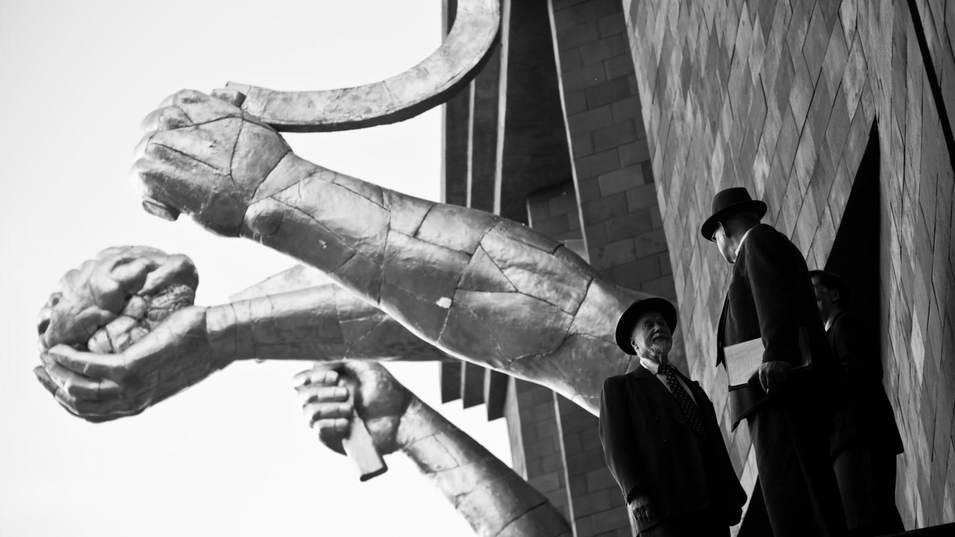 Zwei Männer in Anzügen stehen vor einer an sowjetische Propaganda-Ästhetik erinnernder Skulptur