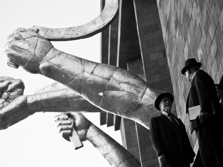 Zwei Männer in Anzügen stehen vor einer an sowjetische Propaganda-Ästhetik erinnernder Skulptur