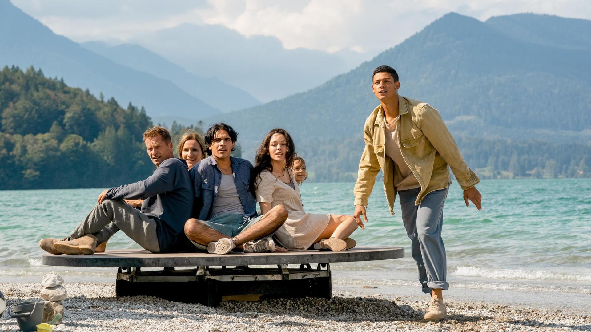 Szenenbild aus "Die Rettung der uns bekannten Welt". Eine Personengruppe ist mit einem Floß am Ufer gestrandet. Im Hintergrund ist ein Bergpanorama mit See zu sehen. Szenenbild aus "Die Rettung der uns bekannten Welt". Eine Personengruppe ist mit einem Floß am Ufer gestrandet. Im Hintergrund ist ein Bergpanorama mit See zu sehen.