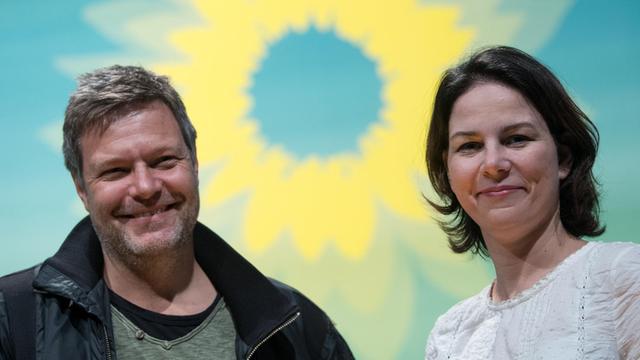 Annalena Baerbock und Robert Habeck stehen vor einem Hintergrund mit einer großen Sonneblumenblume und lachen in die Kamera.