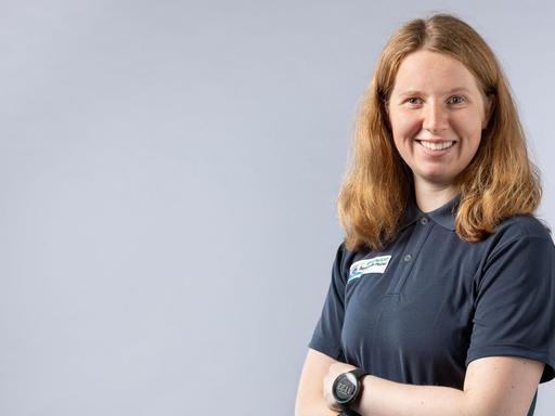 Clara Klug trägt ein graues Poloshirt, steht vor einem hellgrauen Hintergrund und lächelt in die Kamera.