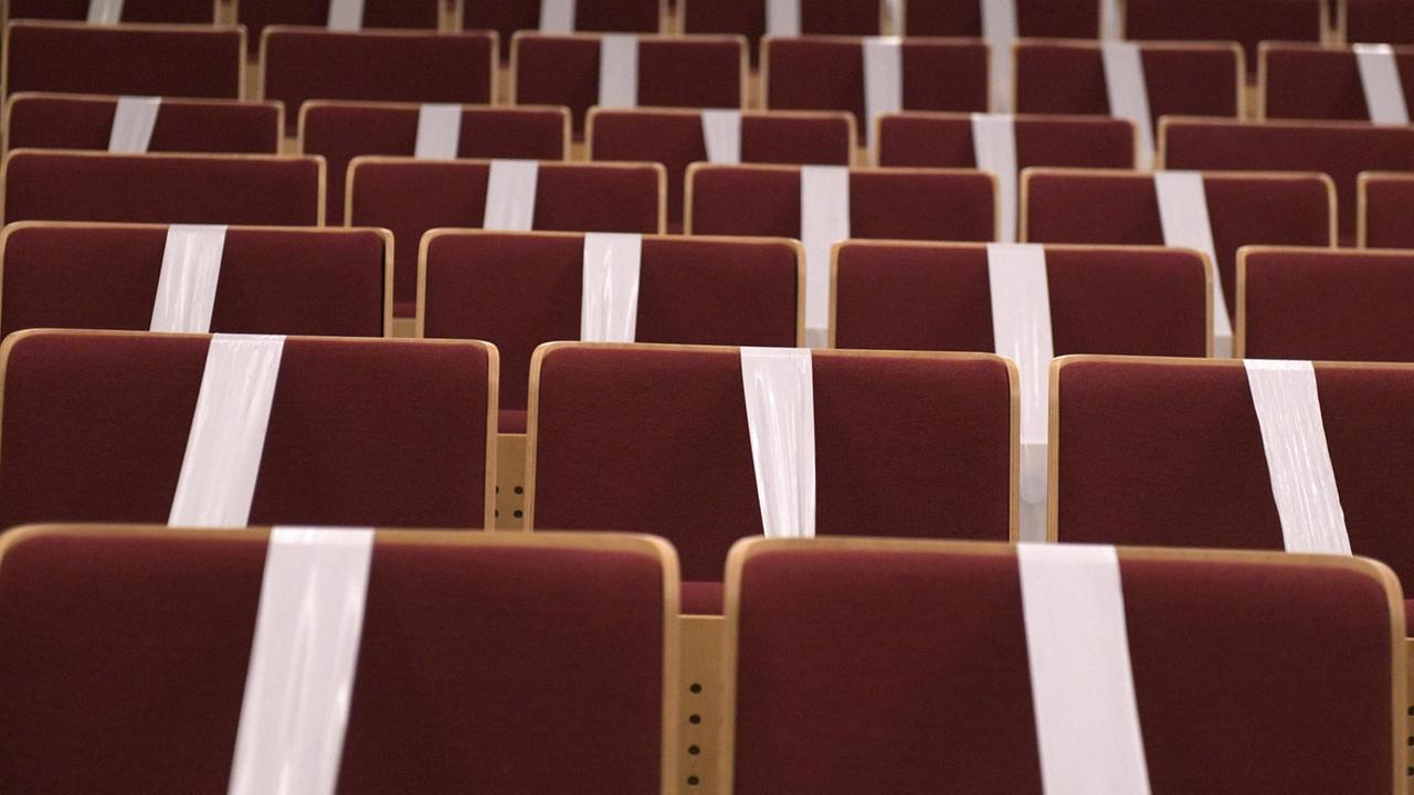 Rote Klappsessel in einem Konzertsaal sind mit weißlichem Tape abgeklebt, sodass sie nicht besetzt werden können. Rote Klappsessel in einem Konzertsaal sind mit weißlichem Tape abgeklebt, sodass sie nicht besetzt werden können.