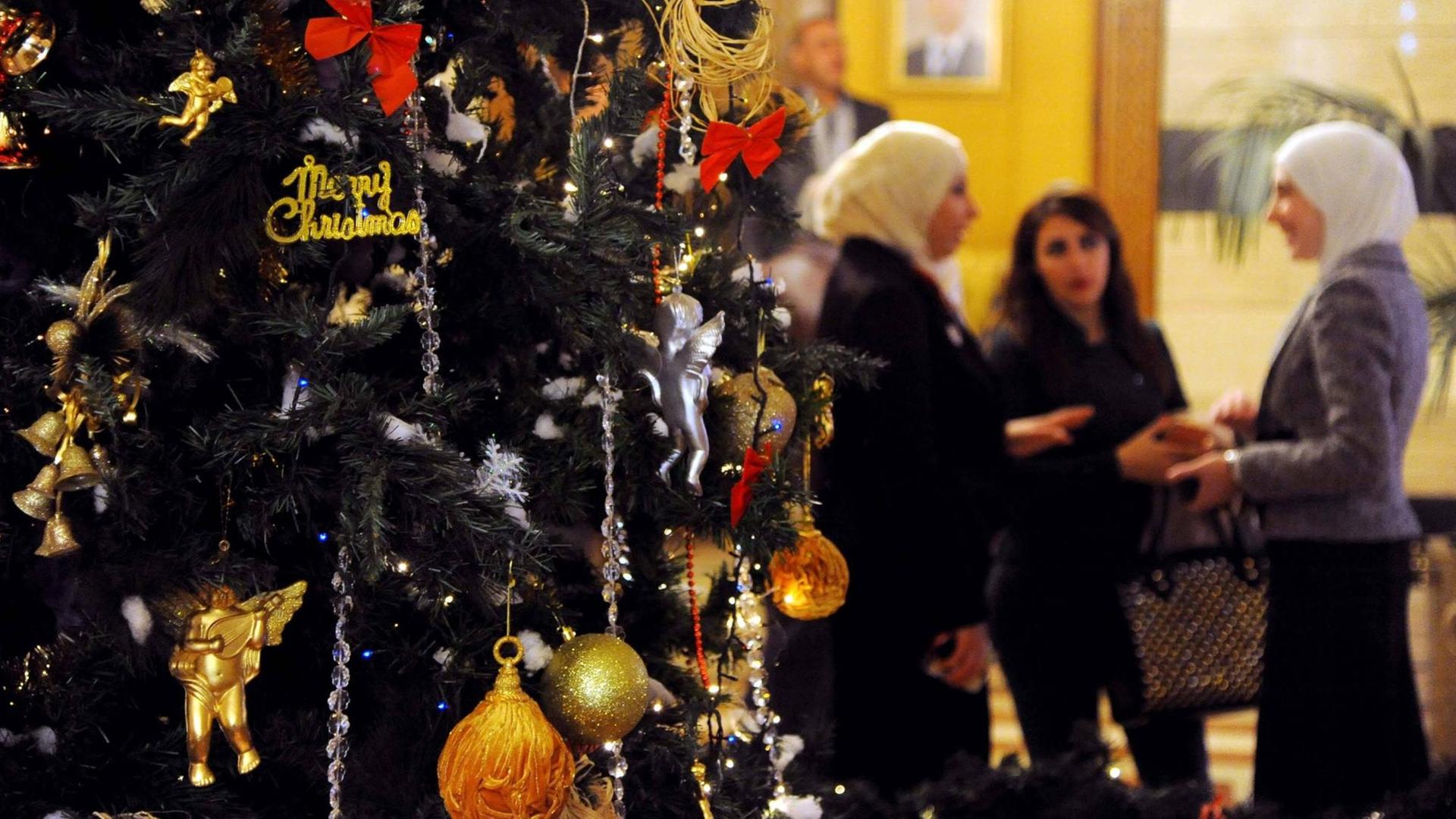 Ein Weihnachtsbaum im syrischen Damaskus. Im Hintergrund sieht man drei Frauen, die sich unterhalten. Zwei von ihnen tragen weiße Kopftücher. Ein Weihnachtsbaum im syrischen Damaskus. Im Hintergrund sieht man drei Frauen, die sich unterhalten. Zwei von ihnen tragen weiße Kopftücher.