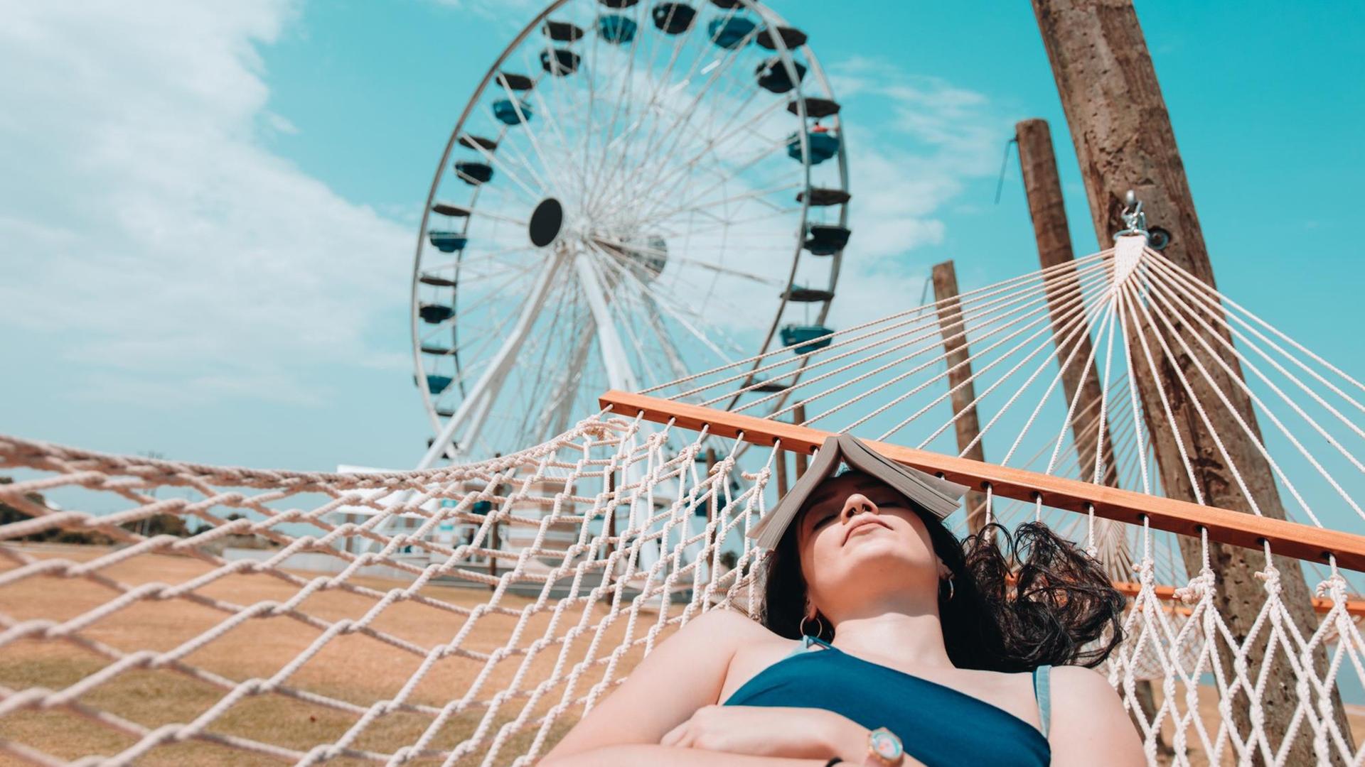 Eine junge Frau schläft in einer Hängematte und hat sich ein Buch aufs Gesicht gelegt. Im Hintergrund ist ein Riesenrad zu sehen. Eine junge Frau schläft in einer Hängematte und hat sich ein Buch aufs Gesicht gelegt. Im Hintergrund ist ein Riesenrad zu sehen.