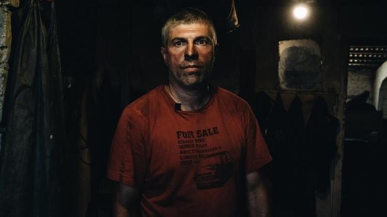 Wadym steht mit verrußtem roten Shirt in einem dunklen Raum und sieht eher resigniert aus. Wadym steht mit verrußtem roten Shirt in einem dunklen Raum und sieht eher resigniert aus.