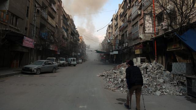 Ost-Ghouta: Ein Mann läuft in der Stadt Douma eine mit Trümmern übersäte Straße entlang, im Hintergrund sind Rauchwollken zu sehen.