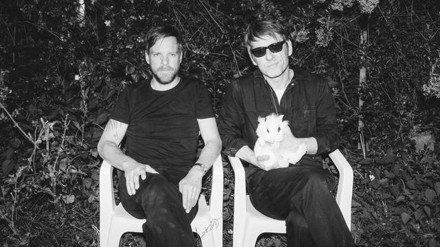 Zwei Männer sitzen mit überschlagenen Beinen auf Gartenstühlen. Einer von ihnen trägt eine dunkle Sonnenbrille und hält ein weißes Kaninchen in den Händen