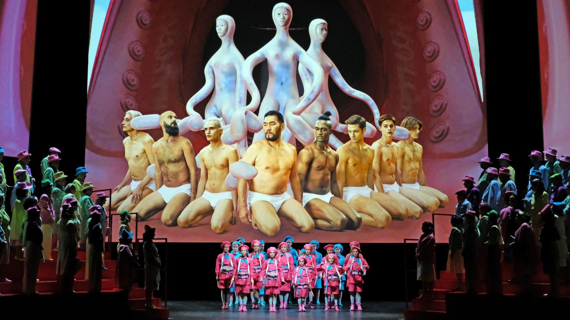 Szenenfoto: Rechts und links ein bunt gekleideter Chor, in der Mitte ein bunt gekleideter Jugend-Chor. Im Hintergrund eine riesige Projektion von acht Männern in weißen Unterhosen, hinter ihnen: drei nicht-menschliche Figuren mit langen Armen.
