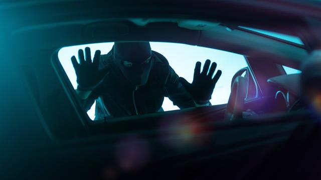 Ein Mann mit schwarzer Maske und Handschuhen steht bedrohlich an einem Autofenster.