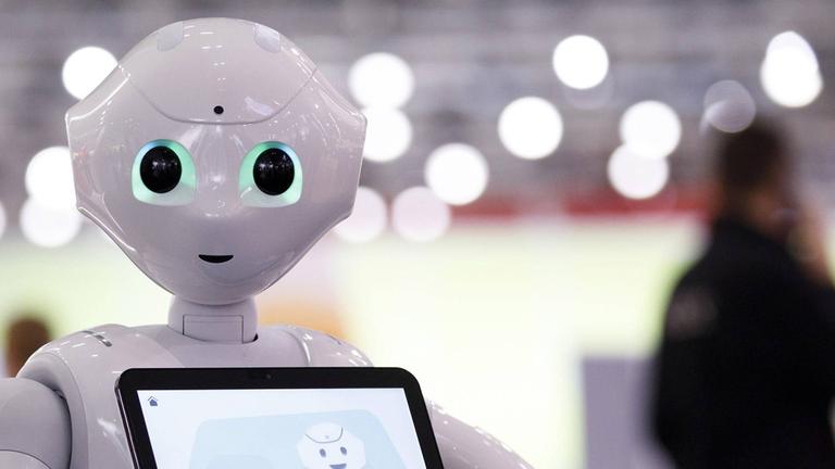 Ein Roboter mit einem Tabelet steht in einer Messehalle und begrüßt die Passanten. Ein Roboter mit einem Tabelet steht in einer Messehalle und begrüßt die Passanten.