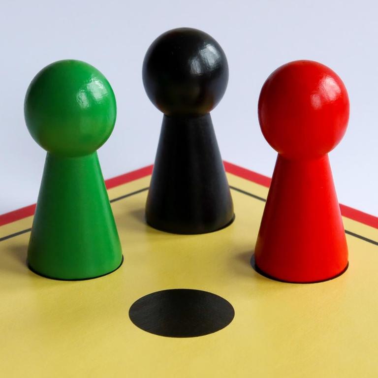 Auf dem Spielfeld von "Mensch ärgere dich nicht" stehen drei Spielfiguren in den Farben Schwarz, Rot und Grün.
