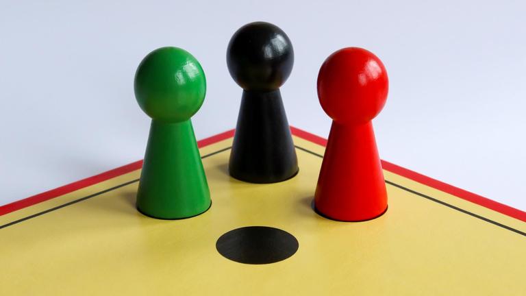 Auf dem Spielfeld von "Mensch ärgere dich nicht" stehen drei Spielfiguren in den Farben Schwarz, Rot und Grün.