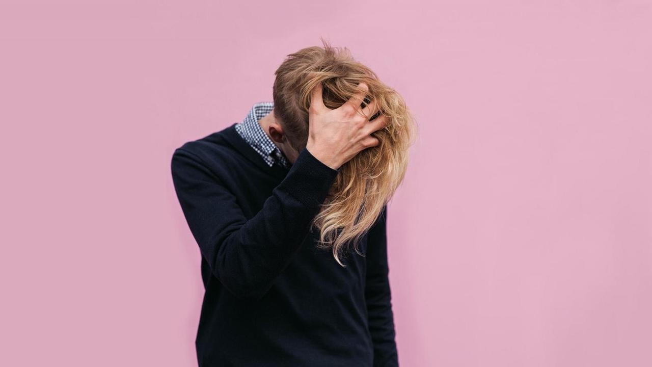 Ein junger Mann vor einem rosa Hintergrund greift sich an den Kopf, der halbrasiert ist und auf der anderen Seite noch sehr lange Haare hat. Ein junger Mann vor einem rosa Hintergrund greift sich an den Kopf, der halbrasiert ist und auf der anderen Seite noch sehr lange Haare hat.
