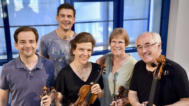Zwei Musikerinnen und drei Musiker stehen mit ihren Streichinstrumenten vor einer verglasten Wand und lächeln in die Kamera.