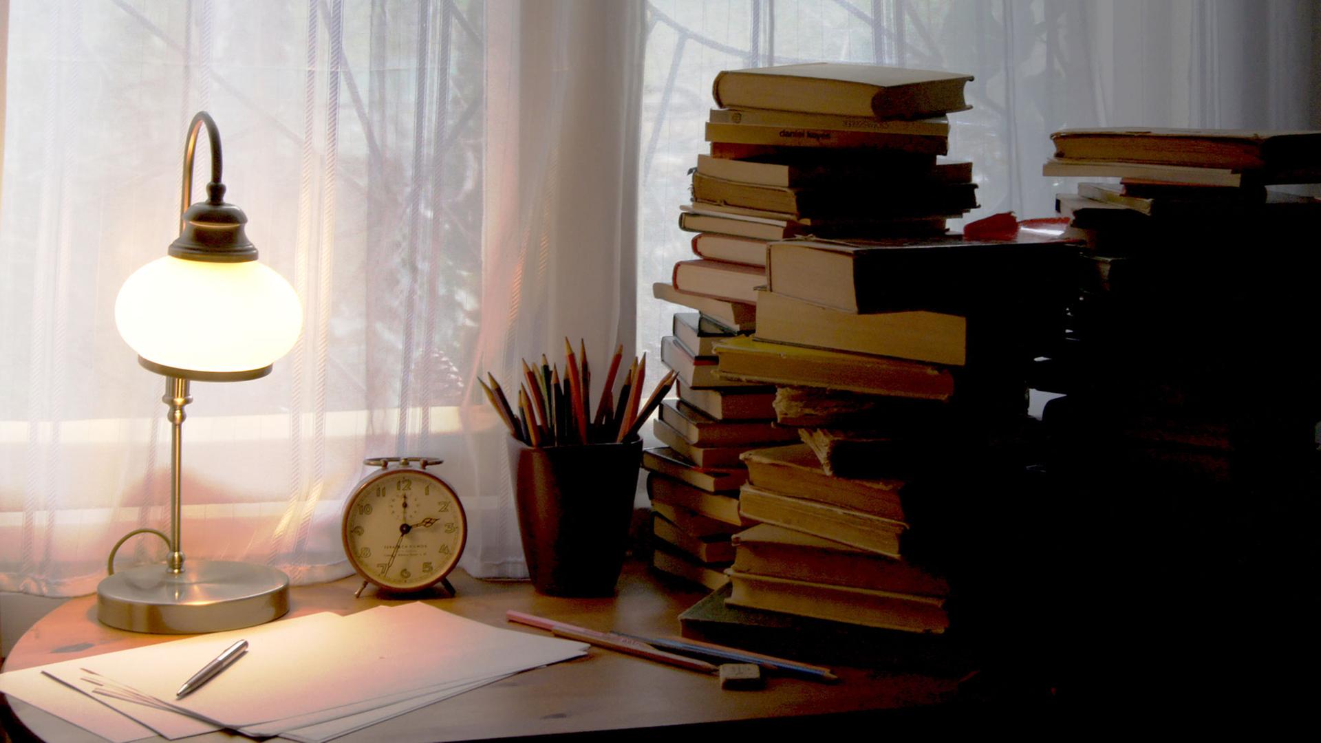 Ein Schreibtisch, mit einer Tischlampe, einem Wecker und gestapelten Büchern.