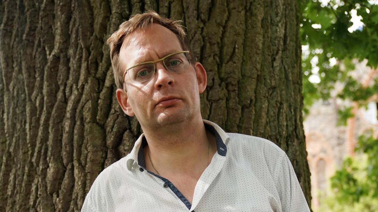 Clemens Meyer porträtiert am Rande einer Lesung auf dem Gut Bülowsiege im August 2019. Er steht im Halbschatten vor einem Baumstamm mit strengem Blick. Clemens Meyer porträtiert am Rande einer Lesung auf dem Gut Bülowsiege im August 2019. Er steht im Halbschatten vor einem Baumstamm mit strengem Blick.
