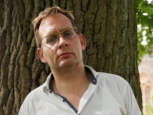 Clemens Meyer porträtiert am Rande einer Lesung auf dem Gut Bülowsiege im August 2019. Er steht im Halbschatten vor einem Baumstamm mit strengem Blick.