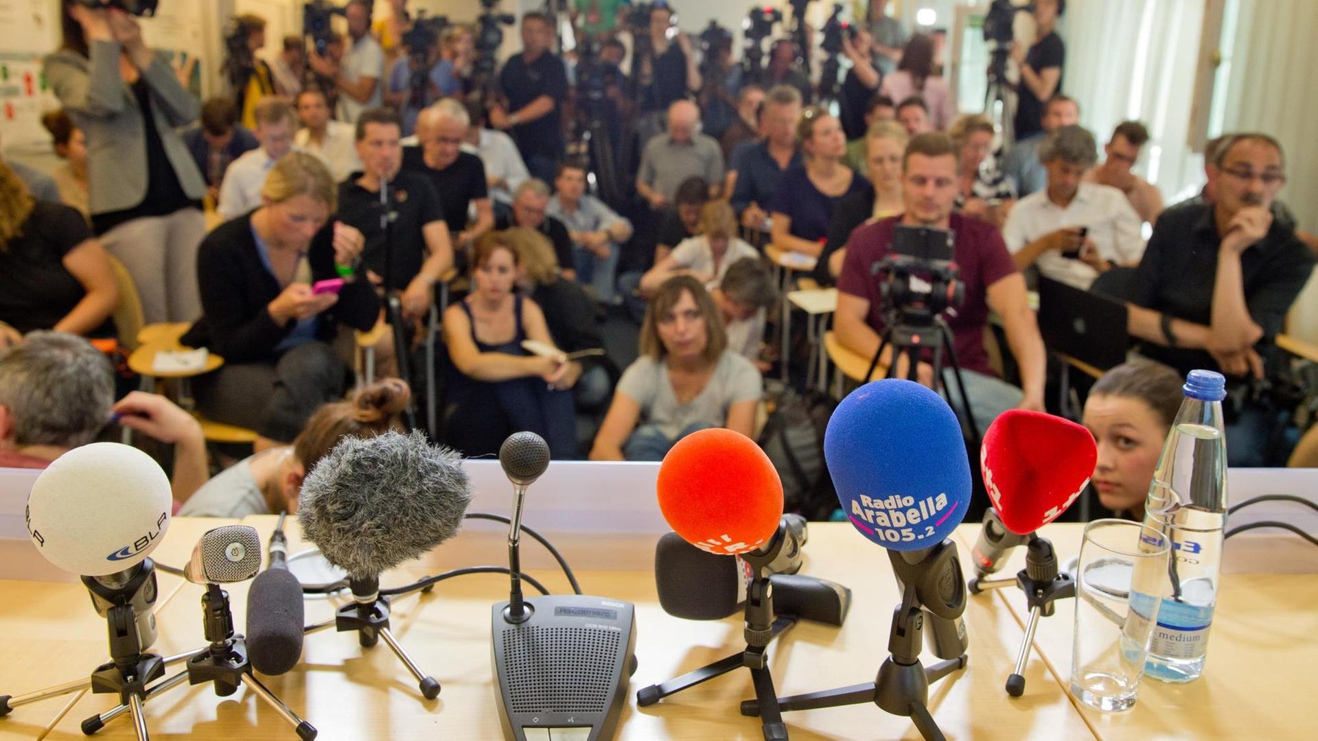 Sie sehen bunte Mikrofone und im Hintergrund viele Journalisten.
