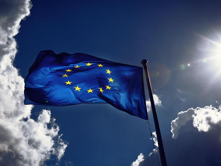 Die Fahne der Europäischen Union weht im Wind und wird von der Sonne angestrahlt. Gewitterwolken ziehen vorbei.