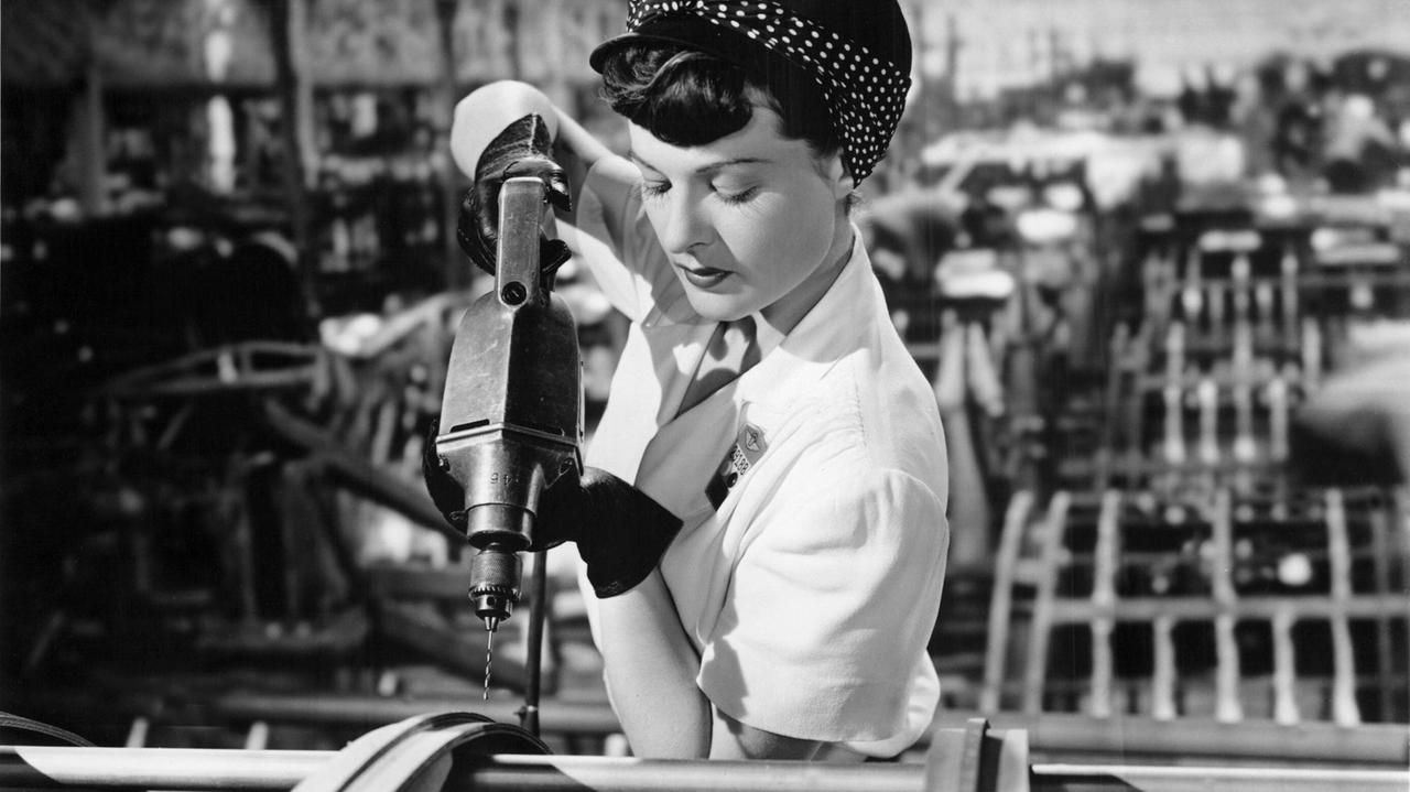 Eine Frau in Mode der 40er-Jahre steht in einer Fabrik und schraubt mit einer Maschine Schrauben ein.
