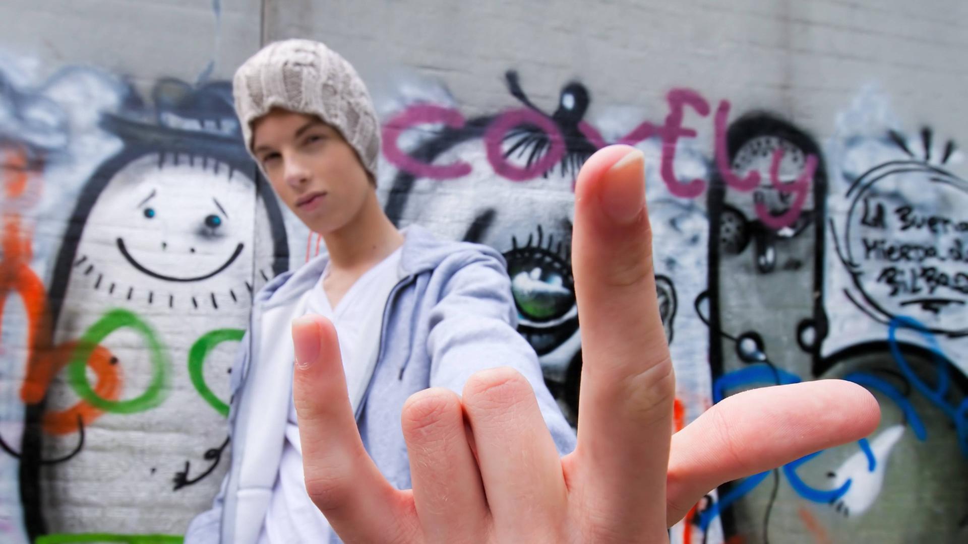 Ein cool blickender jugendlicher Mann gestikuliert vor einer Wand mit Graffiti Ein cool blickender jugendlicher Mann gestikuliert vor einer Wand mit Graffiti