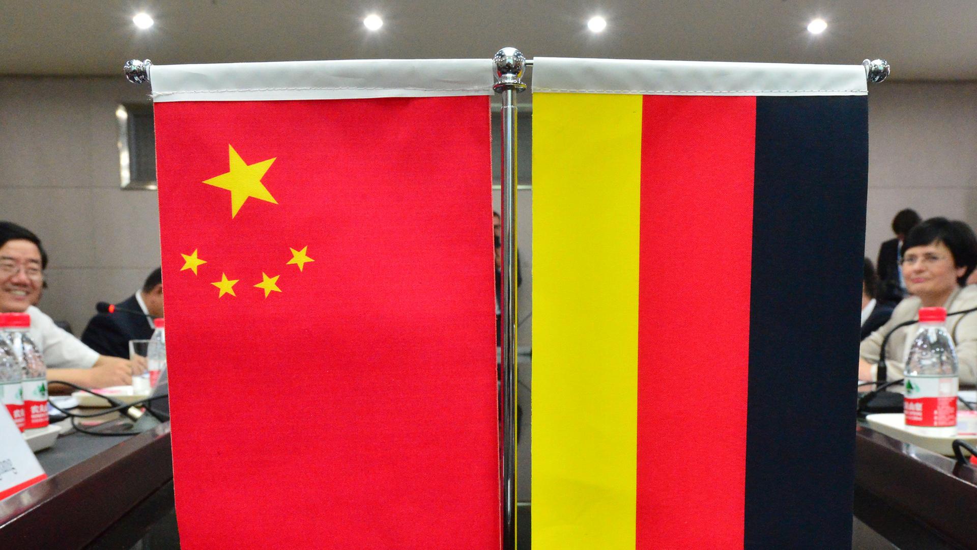 Eine Miniatur-Chinaflagge hängt neben einer Miniatur-Deutschland-Flagge an einem Ständer, der auf einem Tisch steht. Im Hintergrund sitzen Menschen am Tisch. Eine Miniatur-Chinaflagge hängt neben einer Miniatur-Deutschland-Flagge an einem Ständer, der auf einem Tisch steht. Im Hintergrund sitzen Menschen am Tisch.