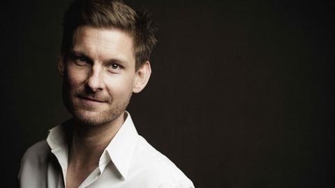 Ein junger Mann mit kurzen braunen Haaren, Dreitagebart und weißem Hemd blickt vor schwarzem Hintergrund freundlich in die Kamera.