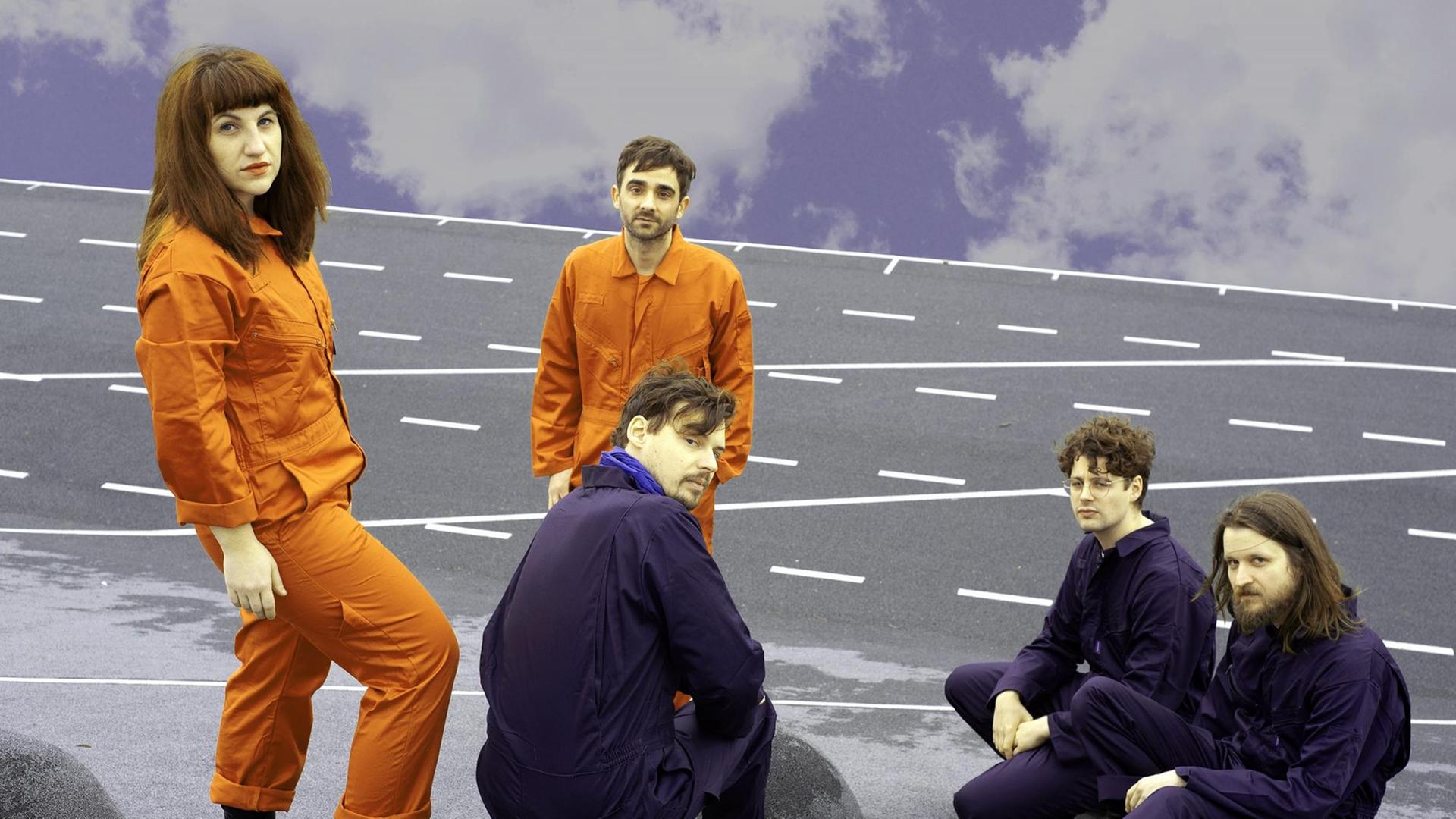 Drei Männer in violetten Overalls sitzen auf einer Fahrbahn. Eine Frau und ein Mann in orangen Overalls stehen daneben.