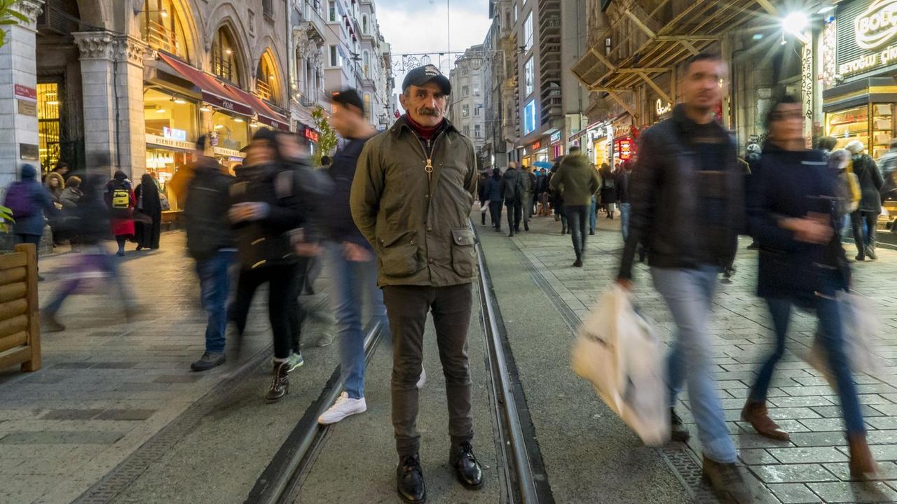 Ein Mann mit dunkler Jacke, Käppi und Schnauzbart steht zwischen den Straßenbahnschienen in einer belebten Fußgängerzone. Ein Mann mit dunkler Jacke, Käppi und Schnauzbart steht zwischen den Straßenbahnschienen in einer belebten Fußgängerzone.