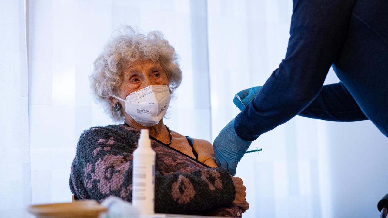 Eine ältere Dame mit grauen Locken und weißem Mundschutz hat den Ausschnitt ihres Pullovers bis über den linken Oberarm gezogen, so dass ein Arzt sie dort impfen kann. Eine ältere Dame mit grauen Locken und weißem Mundschutz hat den Ausschnitt ihres Pullovers bis über den linken Oberarm gezogen, so dass ein Arzt sie dort impfen kann.