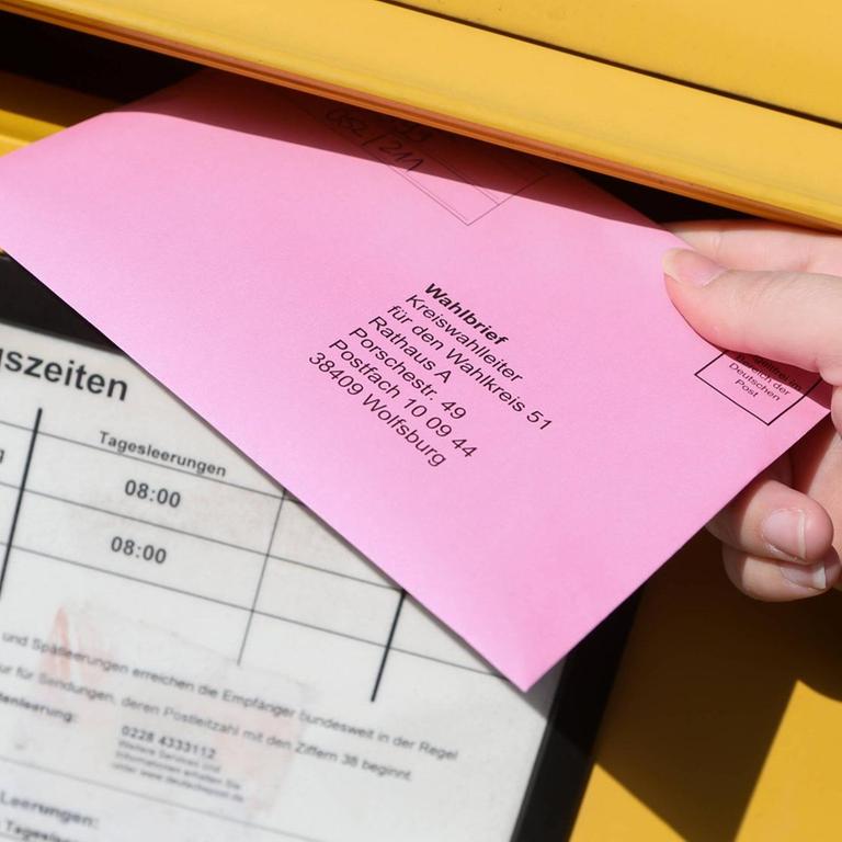 Ein rosafarbener Wahlbrief wird in einen gelben Postkasten eingeworfen.