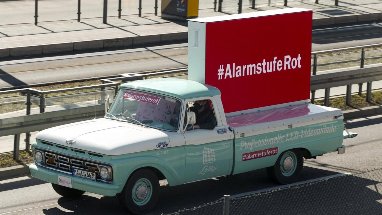 Ein türkisfarbener Laster fährt einen aufgestellten, roten Quader durch eine breite Straße, auf dem der Schriftzug "Alarmstufe rot" zu lesen ist. Ein türkisfarbener Laster fährt einen aufgestellten, roten Quader durch eine breite Straße, auf dem der Schriftzug "Alarmstufe rot" zu lesen ist.