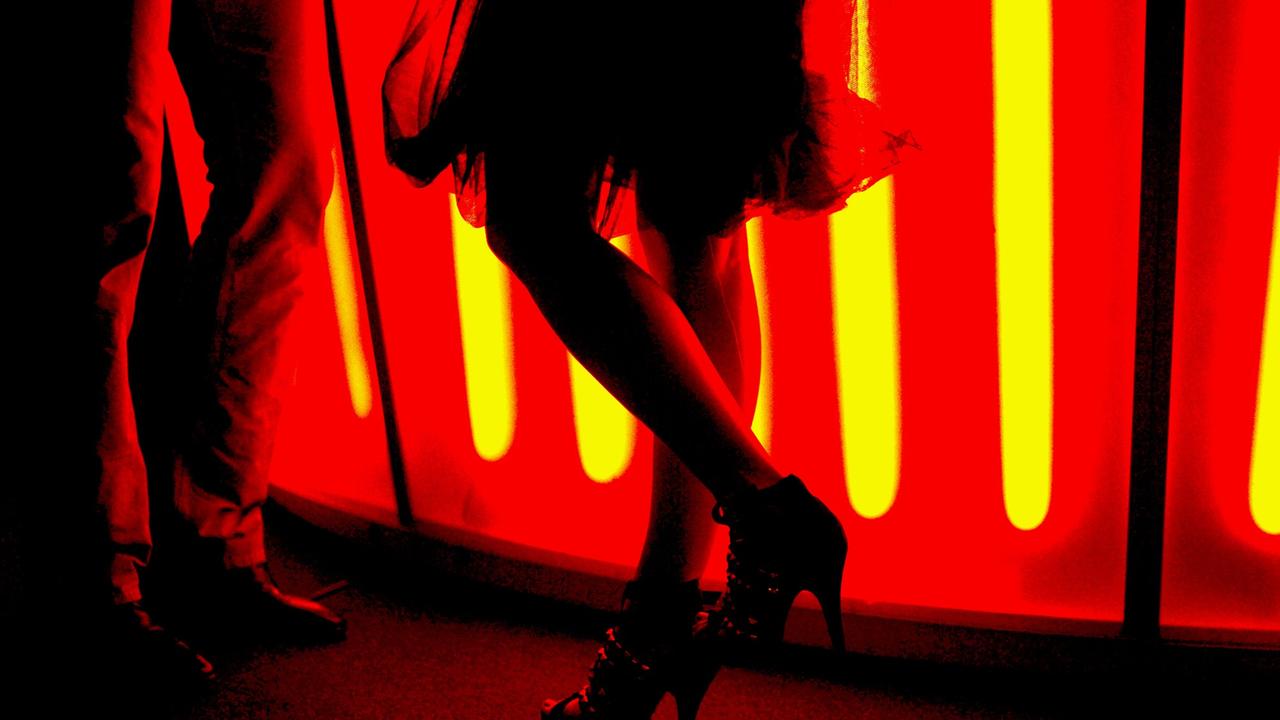Eine Frau mit einem teilweise transparenten Rock steht mit High-Heels an einer rot beleuchteten Bar und unterhält sich mit einem Mann.