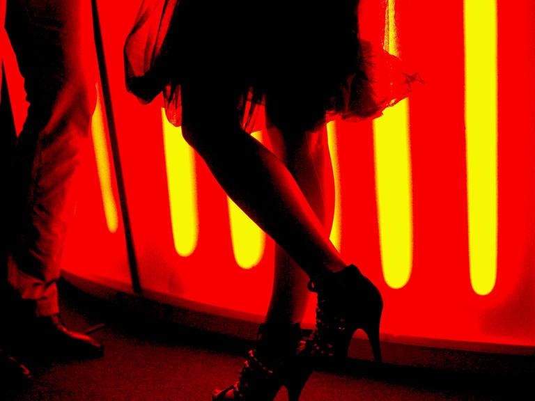 Eine Frau mit einem teilweise transparenten Rock steht mit High-Heels an einer rot beleuchteten Bar und unterhält sich mit einem Mann.