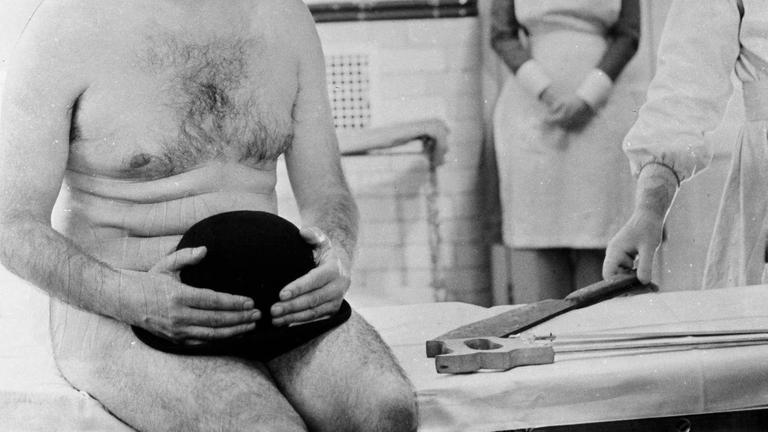 Ein Mann sitzt nackt auf einer Behandlungsliege beim Arzt, nur einen Hut auf dem Schoß (Filmstill aus dem Film "Ulysses", Großbritannien/USA 1967, Regie: Joseph Strick, Darsteller: Milo O Shea)