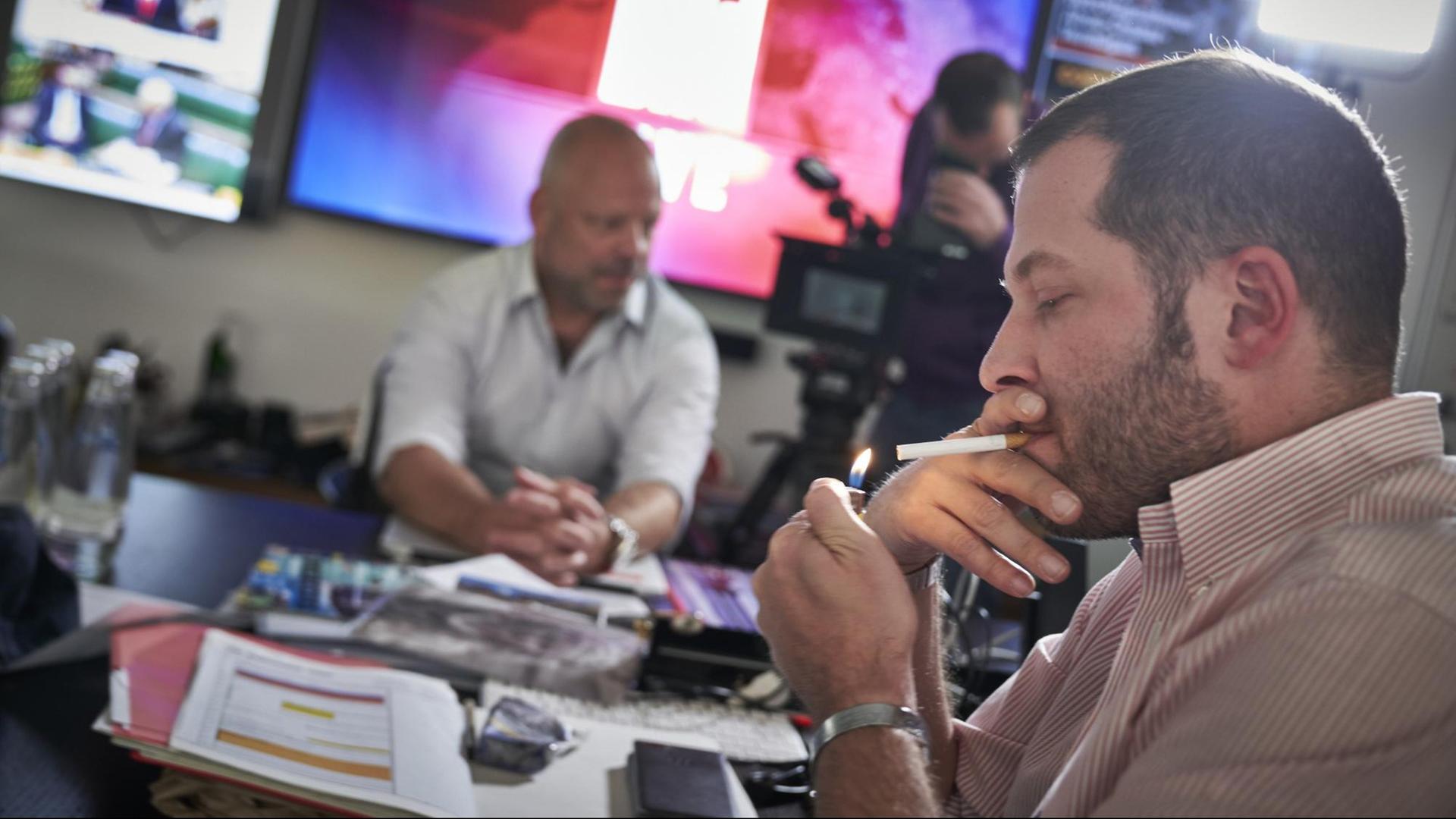 Julian Reichelt, Chefredakteur der Bild-Zeitung, zündet sich eine Zigarette an. Im Hintergrund sitzt ein anderen Mann an einem Tisch, ein weiterer Mann hantiert mit einer Kamera. Julian Reichelt, Chefredakteur der Bild-Zeitung, zündet sich eine Zigarette an. Im Hintergrund sitzt ein anderen Mann an einem Tisch, ein weiterer Mann hantiert mit einer Kamera.
