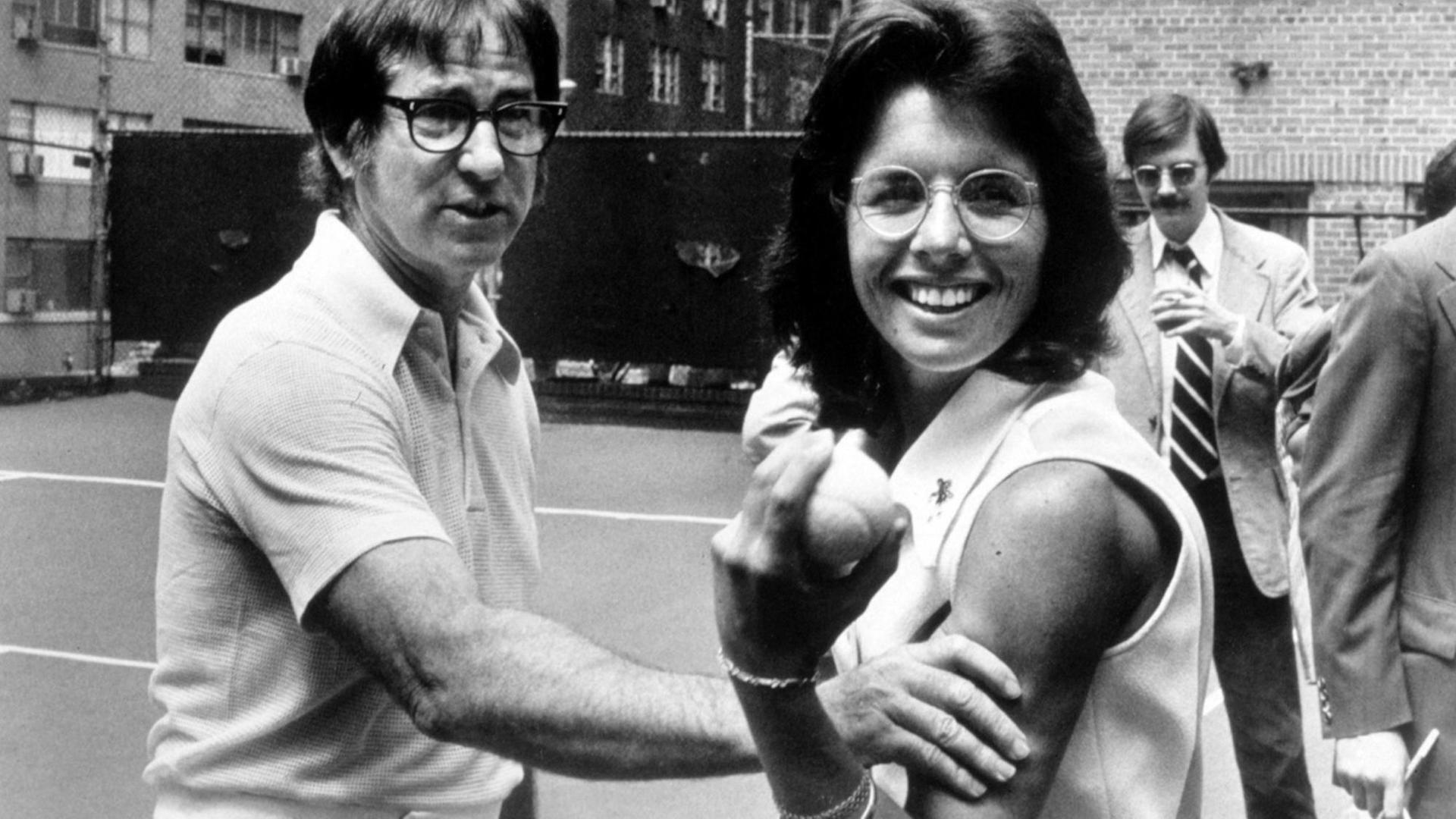 Tennisspieler Bobby Riggs fasst Tennisspielerin Billy Jean King spielerisch an die Armmuskeln, während Billy Jean King mit einem triumphierenden Lachen einen Tennisball in die Kamera hält. Tennisspieler Bobby Riggs fasst Tennisspielerin Billy Jean King spielerisch an die Armmuskeln, während Billy Jean King mit einem triumphierenden Lachen einen Tennisball in die Kamera hält.