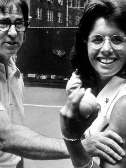 Tennisspieler Bobby Riggs fasst Tennisspielerin Billy Jean King spielerisch an die Armmuskeln, während Billy Jean King mit einem triumphierenden Lachen einen Tennisball in die Kamera hält.