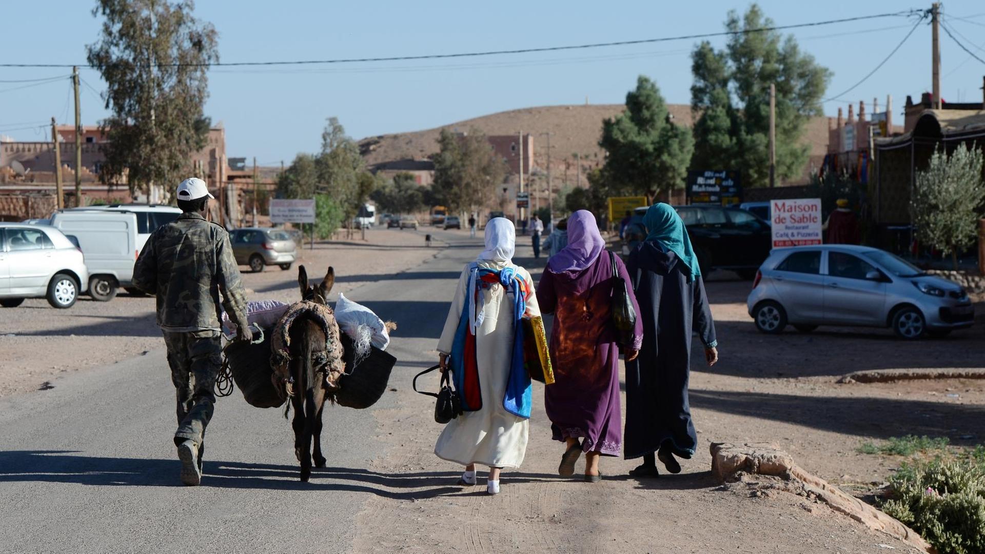  Ein Mann mit einem Esel und drei Frauen in traditioneller Kleidung gehen auf einer Straße in Marokko.