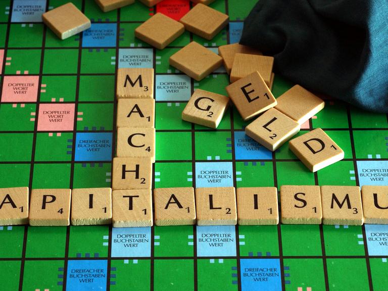 Auf einem Scrabble-Brett sind mit Buchstaben-Steinen die Begriffe "Kapitalismus" und "Macht" zusammengefügt, scheinbar wahllos im Hintergrund liegende Steine bilden das Wort "Geld".