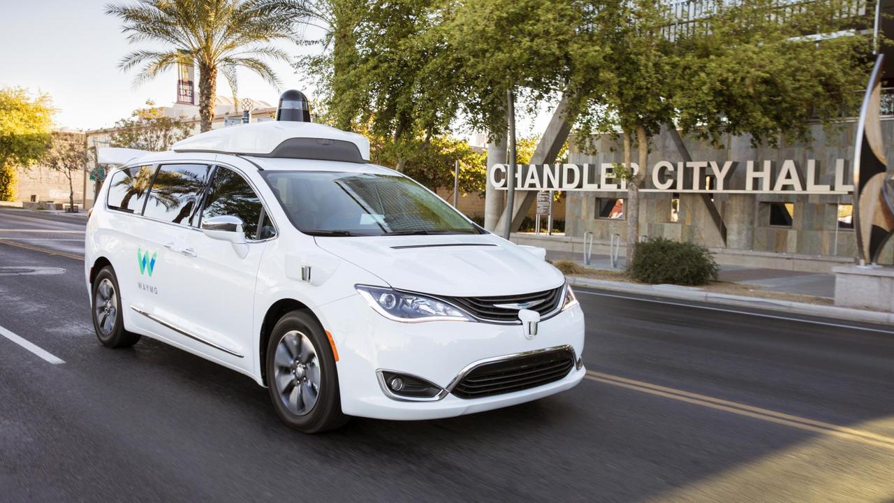 Ein weißer Wagen, beschriftet mit dem Logo von Waymo fährt über die Strasse einer amerikanischen Stadt. Ein weißer Wagen, beschriftet mit dem Logo von Waymo fährt über die Strasse einer amerikanischen Stadt.