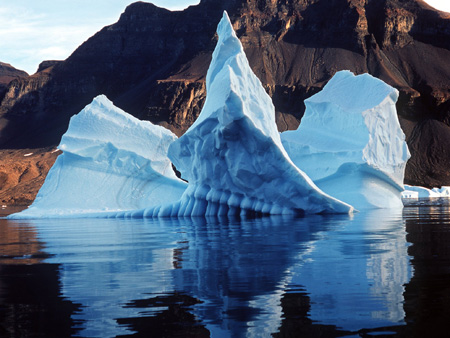 Zu sehen ist ein blauweiß gefärbter Eisberg im grönländischen Meer, dahinter abgeschmolzene Gletscherlandschaft