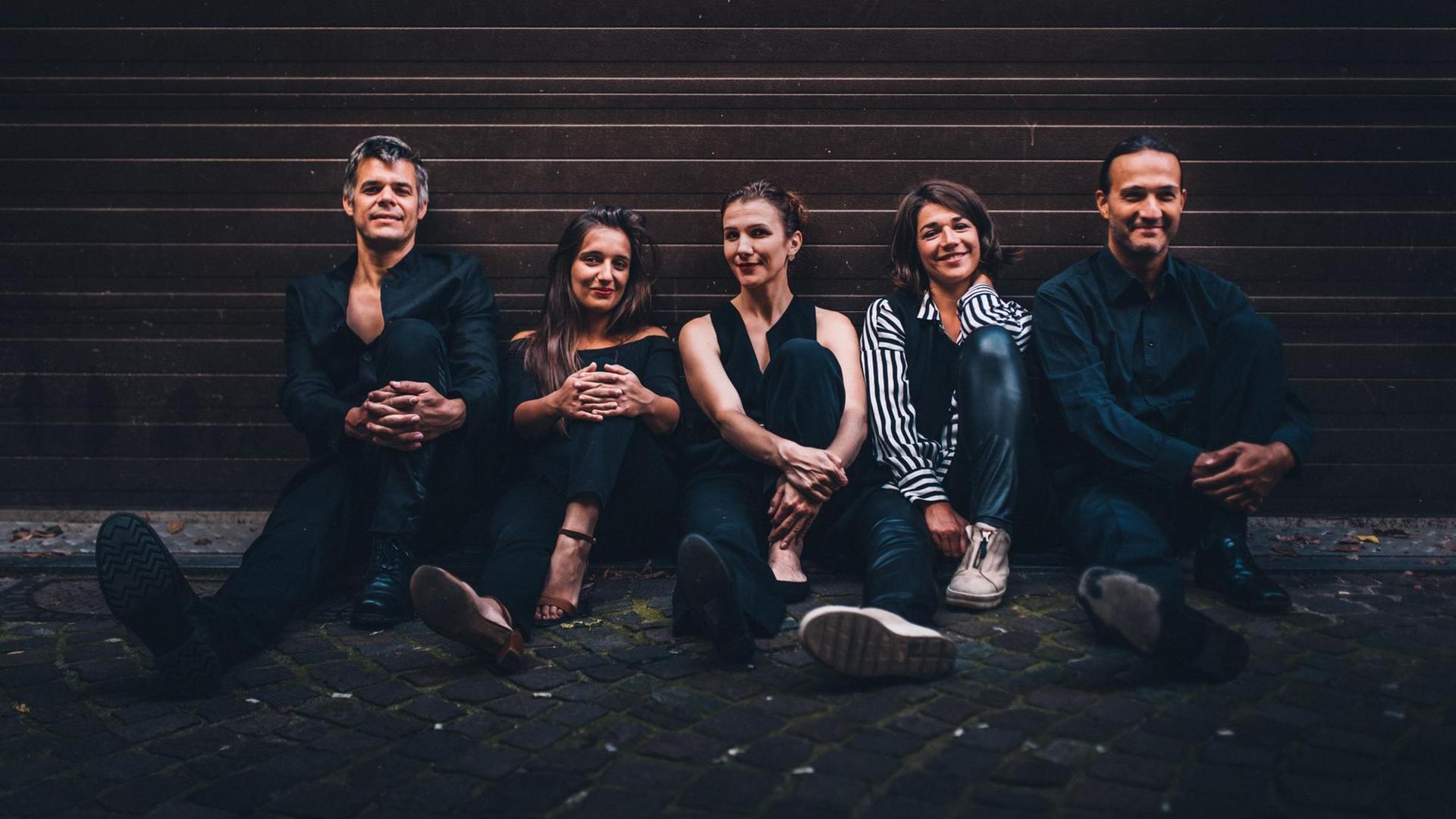 Sieben Musikerinnen und Musiker in legerer Kleidung sitzen nebeneinander vor einer Wand, sie haben alle fröhliche Gesichter. Sieben Musikerinnen und Musiker in legerer Kleidung sitzen nebeneinander vor einer Wand, sie haben alle fröhliche Gesichter.