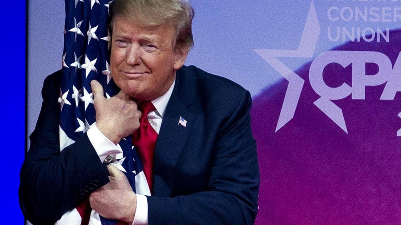 US-Präsident Donald Trump steht auf einer Bühne und umarmt eine amerikanische Staatsflaage. US-Präsident Donald Trump steht auf einer Bühne und umarmt eine amerikanische Staatsflaage.