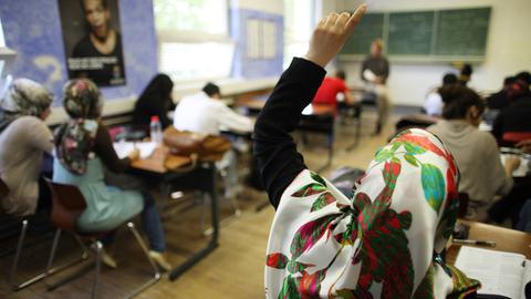 Eine Klasse beim Schulunterricht, die Tische stehen in Zweierreihen, im Hintergrund der Lehrer vor der Tafel, im Vordergrund ein Mädchen mit Kopftuch, das sich meldet.
