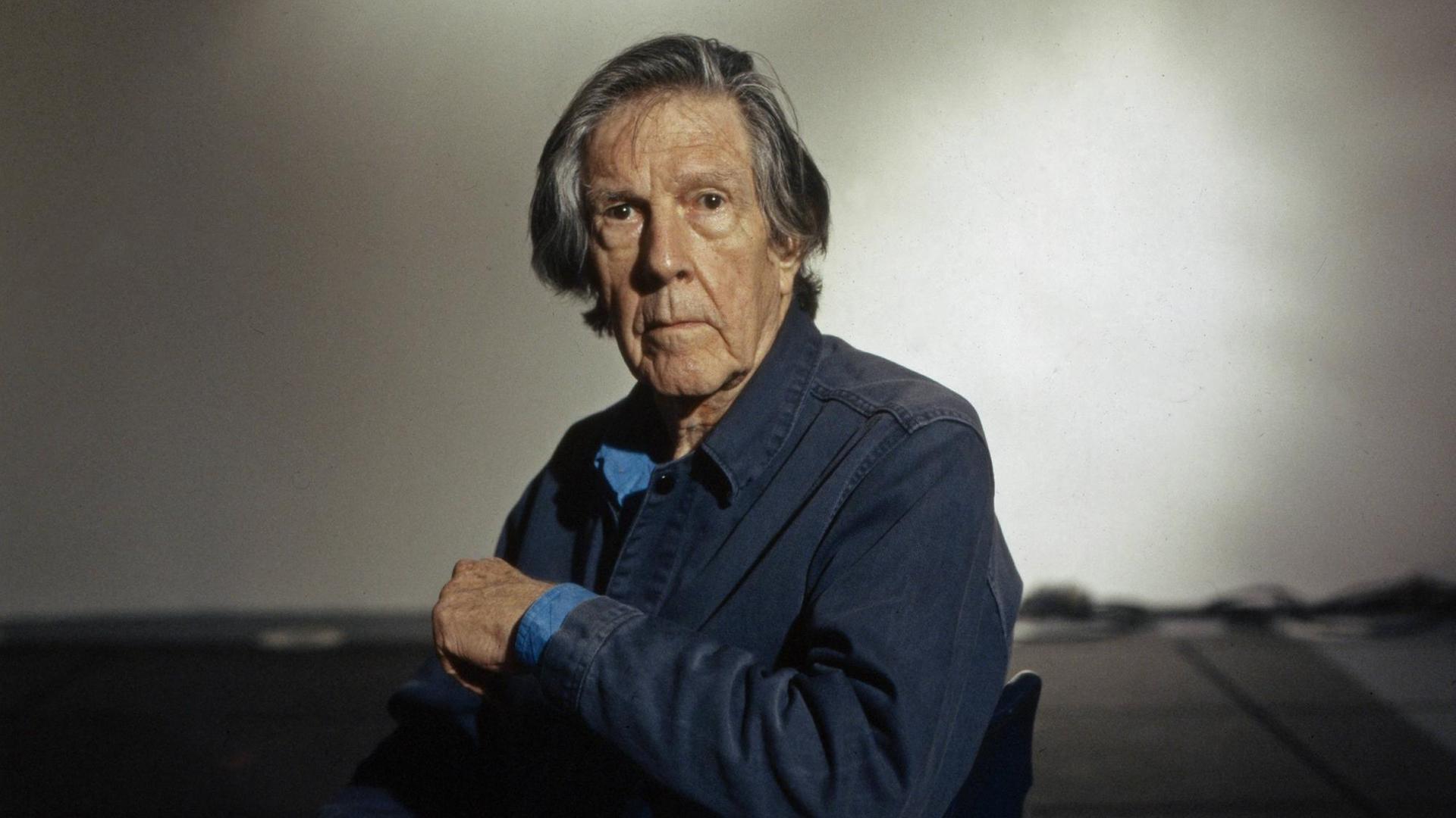 John Cage sitzt seitlich auf einem Stuhl und blickt in die Kamera John Cage sitzt seitlich auf einem Stuhl und blickt in die Kamera