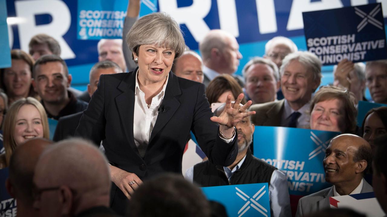 May gestikuliert und spricht, dahinter sitzen vor einem Wahlkampf-Backdrop lächelnde Anhänger. May gestikuliert und spricht, dahinter sitzen vor einem Wahlkampf-Backdrop lächelnde Anhänger.